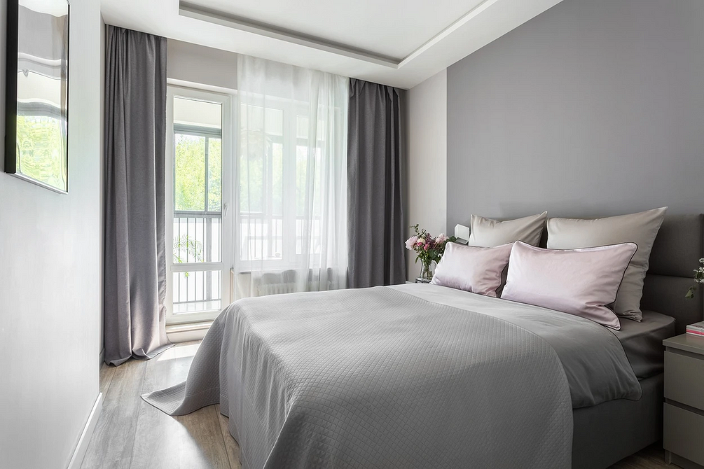 Затишна спальня з сірим ліжком, килимом та декоративними подушками у нейтральних тонах.