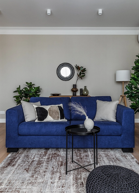 Маленька вітальня з синім диваном, журнальним столиком та круглим дзеркалом.